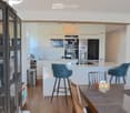 Modern kitchen with white cabinets, stainless steel appliances, and a central island with bar stools. Wooden dining table with chairs and a colorful watermelon-themed centerpiece.