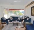 Modern living room with large windows offering a scenic view, featuring a blue sofa, armchairs, coffee tables, a piano, and a fireplace.