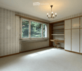 Empty room with white walls, wooden trim, large window, built-in shelving unit, and a small desk with a chair.