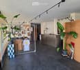 Modern interior with dark flooring, large potted plants, framed artwork, clothing display, and a reception area with a lamp and screen.