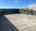 A modern rooftop terrace with a tiled concrete floor, grey planters with greenery, and a low metal railing. The area is clean and spacious, with a clear blue sky in the background.