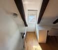 Bright, narrow hallway with wooden flooring, a small desk, and a chair, leading to a window with a view of a roof.