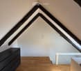 Attic room with wooden floor, slanted wooden beams, white walls, and a small black cabinet and white table.