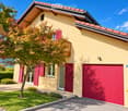 Two-story house with yellow exterior, red shutters, garage, and balcony overlooking a garden area.