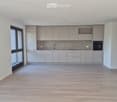 Empty kitchen with light grey cabinets, a sink, and a tiled backsplash. The space features wooden flooring and large windows offering a view of greenery.