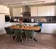 Modern kitchen with white cabinets, wooden dining table, four green chairs, and various appliances including an oven and toaster. The floor is light wood, and the space is well-lit with track lighting.