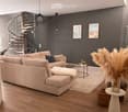 Modern living room with light-colored sofa, coffee table, floor lamp, and decorative items including a vase with dried plants and a small basket. The room features wooden flooring and a dark grey accent wall with framed artwork.