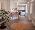 A modern, well-lit hallway leading to a kitchen area. The hallway features a round woven rug on a wooden floor, a small table with a plant and a map, and a chair. The kitchen area has white cabinets and a countertop with various items.