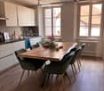 Bright, modern dining area with a large wooden dining table surrounded by green chairs, white kitchen cabinets, and a sink area. Natural light streams in through large windows, and a vase with flowers adds a touch of color.