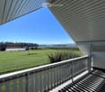 A spacious balcony with white railings and a covered roof, overlooking a green field and distant mountains under a clear blue sky.