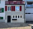 Two-story house with light gray walls, red shutters, and a red balcony railing. The ground floor has a black door and several windows, while the upper floor features additional windows and a small balcony. The roof is tiled with brown tiles and has dormer