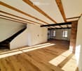 Empty room with wooden floor, exposed wooden beams on the ceiling, white walls, and a staircase leading to another level.