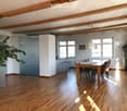 Open-plan living and dining area with wooden floors, exposed wooden beams, large windows, a dining table with chairs, bar stools, and pendant lighting.