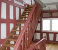 A wooden staircase with red-painted railings and white paneling on the walls, leading up to a higher level. The hallway has wooden flooring and a chandelier hanging from the ceiling.