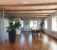 Open-plan dining area with wooden flooring, exposed wooden beams, large windows, dining table with chairs, and a tall potted plant.