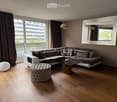 Modern living room with a sectional sofa, coffee table, ottoman, floor lamp, and large mirror. The room features wooden flooring and large windows with curtains.