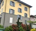 Two-story house with yellow facade, grey shutters, and multiple windows. The house has a well-maintained garden with various shrubs and trees in front.