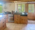 Bright kitchen with wooden cabinets, stainless steel appliances, and a large sink. Features a tiled floor, natural light from windows, and decorative elements like a flower vase and hanging herbs.