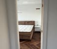 A furnished bedroom with a wooden bed, mattress, and a small nightstand. The room features wooden flooring and white walls.