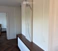 A narrow hallway with wooden flooring, white walls, and a long wooden bench with white drawers. The hallway features several doors and a visible electrical outlet on the wall.