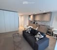 Modern open-plan living and dining area with a grey sofa, coffee table, dining table, chairs, and a small kitchen area with cabinets, sink, and appliances.