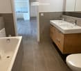 Modern bathroom with a bathtub, double sink, and toilet. The space features wooden cabinets, tiled walls, and a view into an adjacent kitchen area.