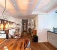 Open-plan living and dining area with wooden flooring, a large wooden dining table with chairs, a cat tree, a modern kitchen area with cabinets and appliances, and a hanging light fixture with exposed bulbs.