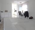 Bright, modern dining area with a white dining table and six chairs, a small kitchen area with white cabinets and appliances, and decorative elements including a vase with flowers and a suitcase on the floor.