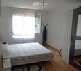 Bedroom with a double bed, nightstand, mirror, and ironing board. Light-colored walls, wooden flooring, and a window with curtains.