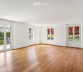 Empty room with light wooden flooring, white walls, and large windows with views of greenery outside. The space is unfurnished and well-lit.