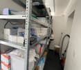 A storage room with metal shelving units filled with various boxes and items, a small white stool, and a vacuum cleaner leaning against the wall.