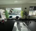 Bright, modern office space with large windows, a desk, chairs, a laptop, a fan, and potted plants.