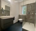 Modern bathroom with a dark blue floor, white sink, mirror, and a glass-enclosed shower area. Includes a toilet, towel, and toothbrush holders.