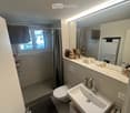 Modern bathroom with a white sink, chrome faucet, toilet, large mirror, and a shower area with a window and curtain. The countertop holds various toiletries and decorative items.