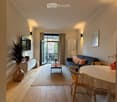 Bright living room with wooden flooring, a sofa, coffee table, dining table, and chairs, with a large window leading to a balcony.