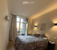 Modern bedroom with a large bed, nightstand, wall-mounted shelf, and a view through an open window to a balcony area.