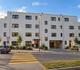 Modern two-story building with white facade, multiple windows, and a covered entrance. There is a parking area on the right side and a pedestrian crossing in front.