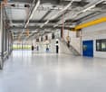 Wide, empty industrial-style corridor with grey concrete floor, exposed metal beams, and hanging orange safety equipment. Features large windows, blue doors, and a staircase leading to an upper level.