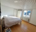 Bright bedroom with wooden flooring, slanted ceiling, large window offering a view, bed with white bedding, nightstand with books, and a wardrobe.