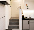 Modern hallway with white walls, grey carpet, a small bench, and a staircase leading to an upper level. A small kitchen area is visible with a sink, faucet, and countertop.