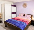 A modern bedroom with a wooden bed, colorful bedding, a nightstand with a lamp, a vase with dried flowers, and a large wardrobe with a purple accent.