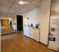 Reception area with a white reception desk, brochures, a chair, a small table, and a potted plant. The floor is wooden, and the ceiling has recessed lighting.