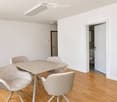 Bright, minimalist room with light wooden flooring, white walls, and modern furniture including a dining table with four chairs and a cabinet.