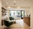 Modern, well-lit room with wooden flooring, large windows, green armchairs, a round coffee table, a reception desk, and shelves with books and decorative items.
