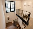 Modern staircase with wooden steps, black metal railing, and integrated lighting. Features a large window, framed artwork, and a fire extinguisher mounted on the wall.