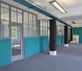 Modern corridor with teal and white walls, large windows, grey carpeted floor, and visible pillars.
