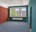 Empty room with grey carpet, teal and red walls, large windows with blinds, radiator, and a partially visible adjacent space.