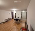 A furnished room with wooden flooring, a table with chairs, a TV screen, and a remote control on a small table. There are boxes and storage items near the door.