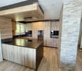 Modern kitchen with wooden cabinets, a central island, and a stone accent wall. Features include a black countertop, a sink, and a window providing natural light.