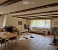 Living room with exposed wooden beams, tiled floor, two cream-colored sofas, a wooden dining table with four chairs, a fireplace with decorative items, a bookshelf, and a television setup.